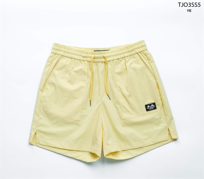 Quần short màu vàng nhạt cạp chun dây rút, hai túi đắp trước, miếng logo nhỏ, xẻ tà gấu, trải phẳng nền trắng