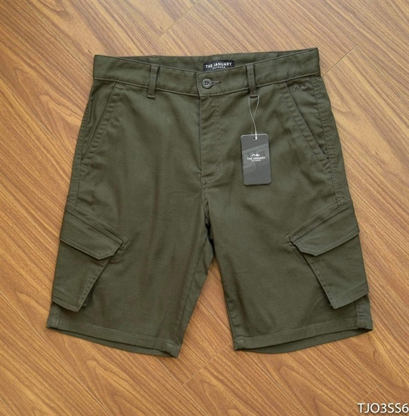 Quần short túi hộp màu xanh rêu trải phẳng trên sàn gỗ, mặt trước, túi hộp nắp gập hai bên, có thẻ treo