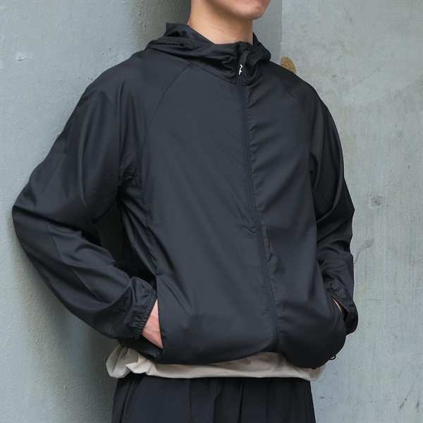Người mẫu mặc áo gió đen có mũ, khoá kéo giữa, tay cho vào túi, đứng dựa tường xám