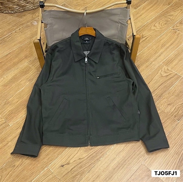 Áo khoác work jacket màu xám than treo móc, cổ bẻ, khoá kéo giữa, túi khoá ngực và hai túi chéo, đặt trên sàn gỗ
