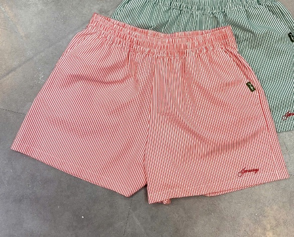 Quần short nữ kẻ sọc hồng trắng cạp chun, chữ thêu ở ống, trải phẳng trên nền xám, phía sau là chiếc sọc xanh lá