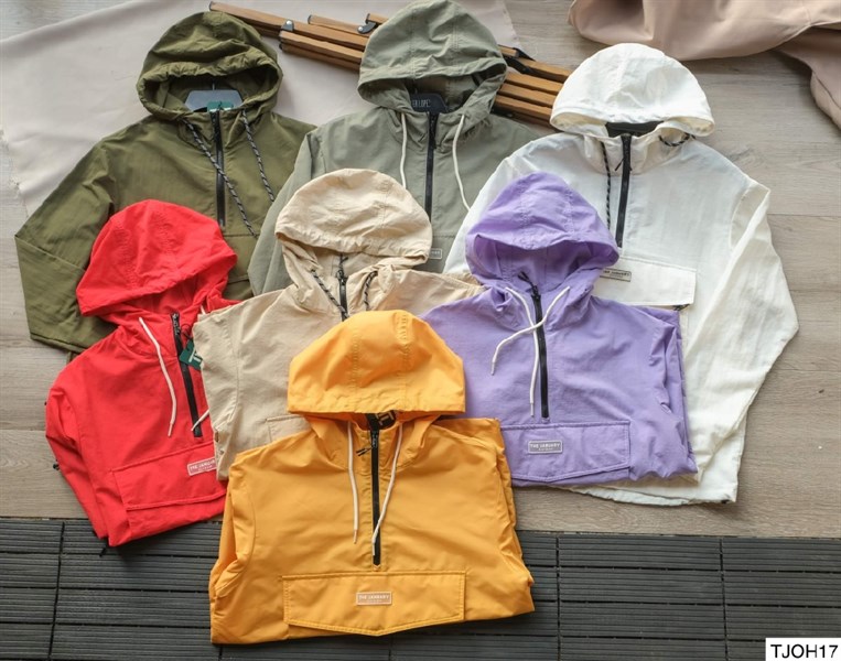 Bảy áo khoác gió chui đầu có mũ xếp chồng trên sàn gỗ: rêu, xanh xám, trắng, đỏ, be, tím, vàng; khoá kéo ngực, túi trước